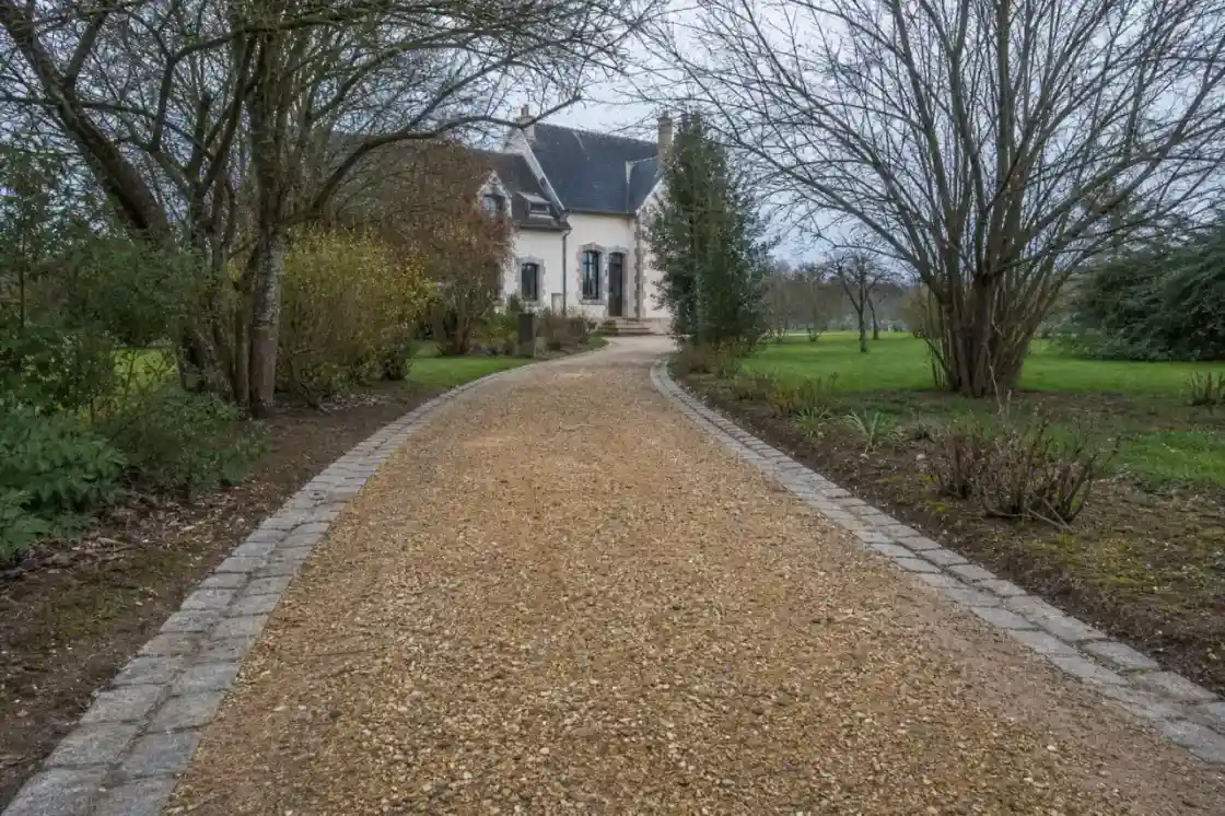 Allée de jardin en gravier stabilisé et pavés menant à une maison de la région castelroussine, sous la lumière basse de la plaine du Berry, sol argilo-calcaire visible en bordure, ambiance calme et humide typique de l'Indre.
