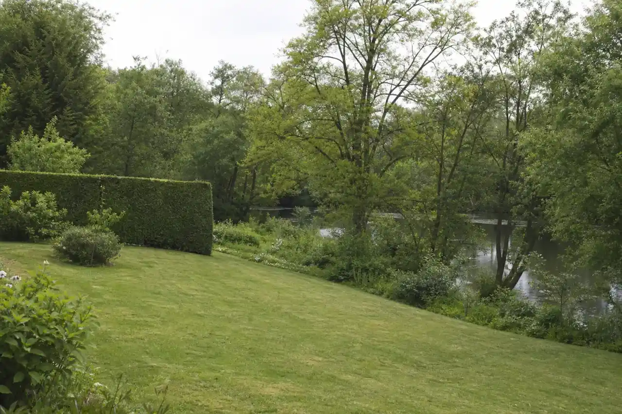 Jardin résidentiel soigné à Déols en fin de matinée de printemps, pelouse rase bordée d'une haie taillée, terrain légèrement en pente vers la végétation fraîche de la rive de l'Indre, lumière douce et basse de plaine berrichonne, cadrage large horizontal.