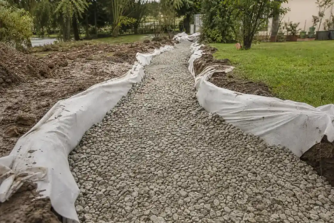 Gros plan sur une fondation drainante en cours de pose, géotextile et grave compactée dans une tranchée d'allée, sur un sol argileux lourd typique de la Champagne berrichonne autour de Châteauroux.