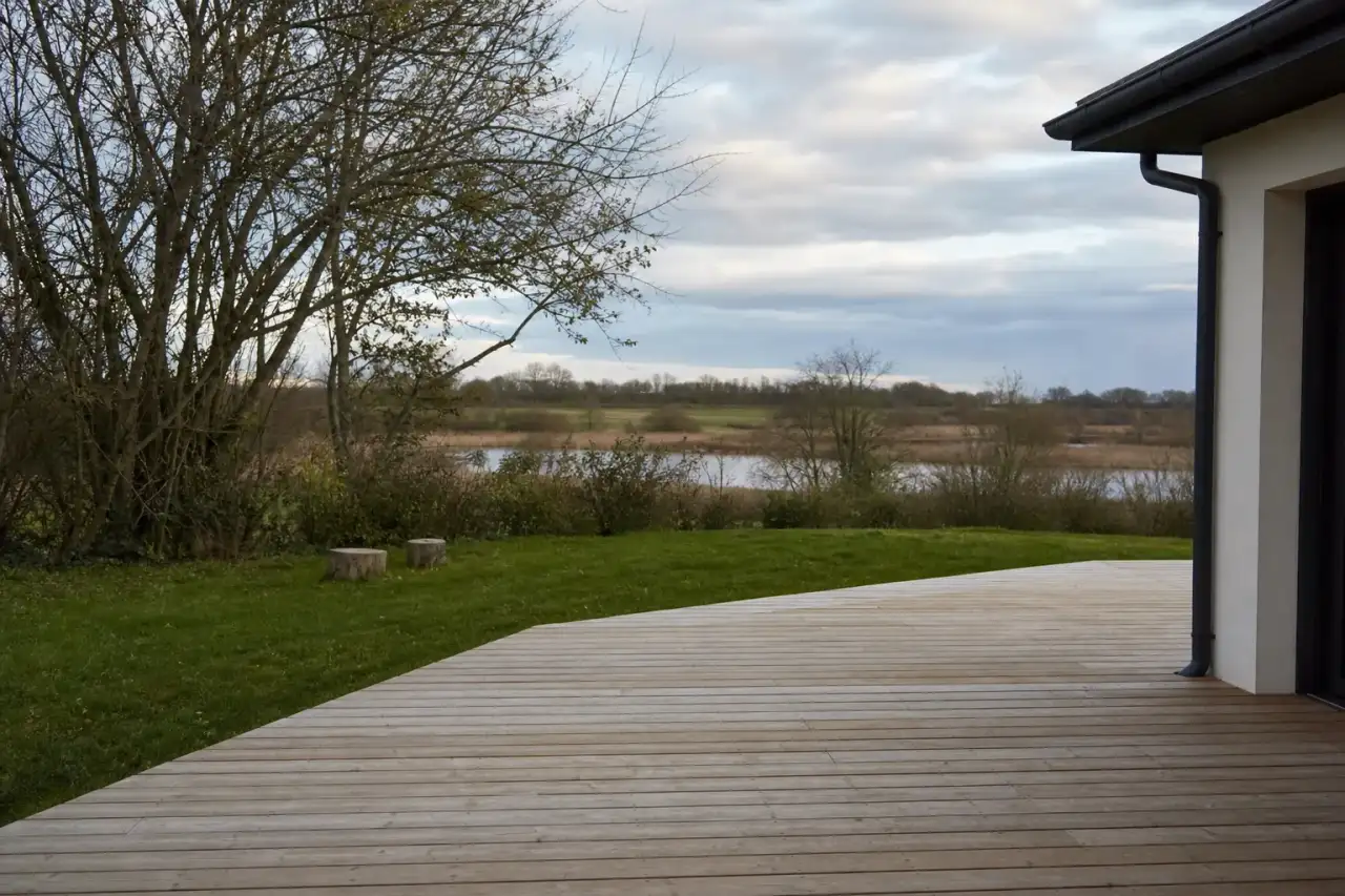 Terrasse en bois clair ouverte sur l'horizon bas de la plaine berrichonne près de Châteauroux, lumière douce de fin d'après-midi, transition nette entre la maison et le jardin, roselières au loin.