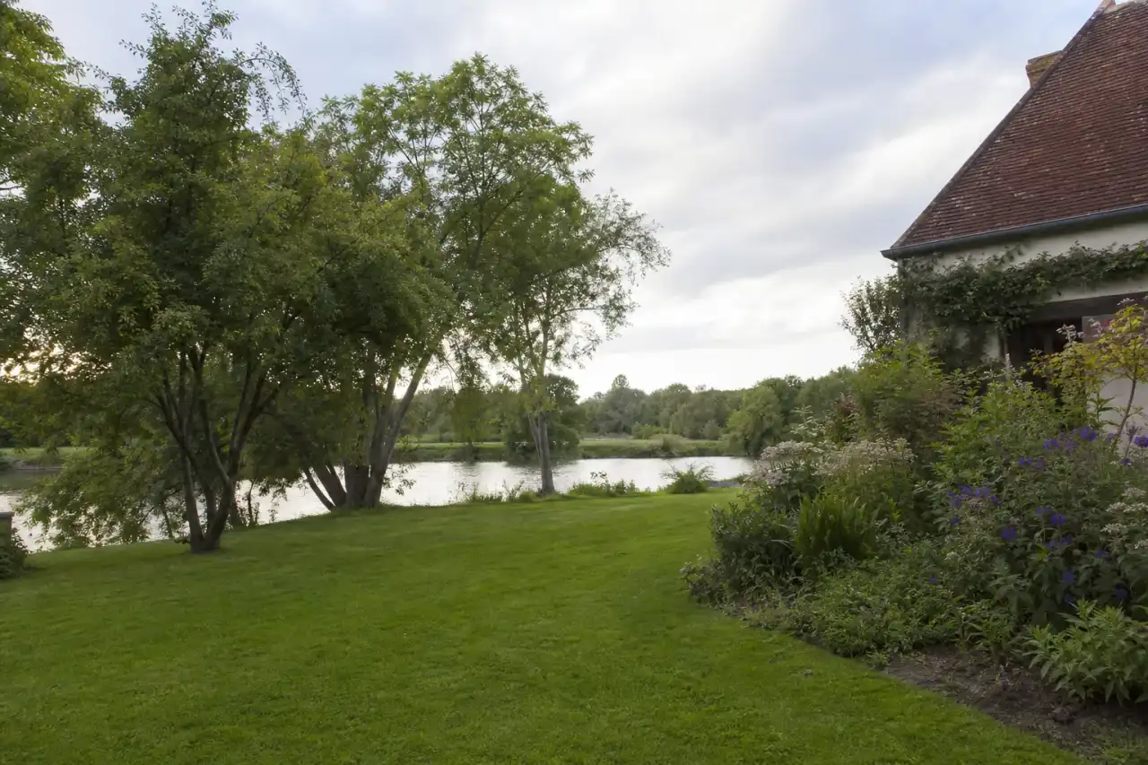 Jardin de pavillon à Saint-Maur en bord d'Indre, pelouse fraîche et massifs verdoyants, lumière douce de fin d'après-midi, cadrage large ouvrant sur la rive.
