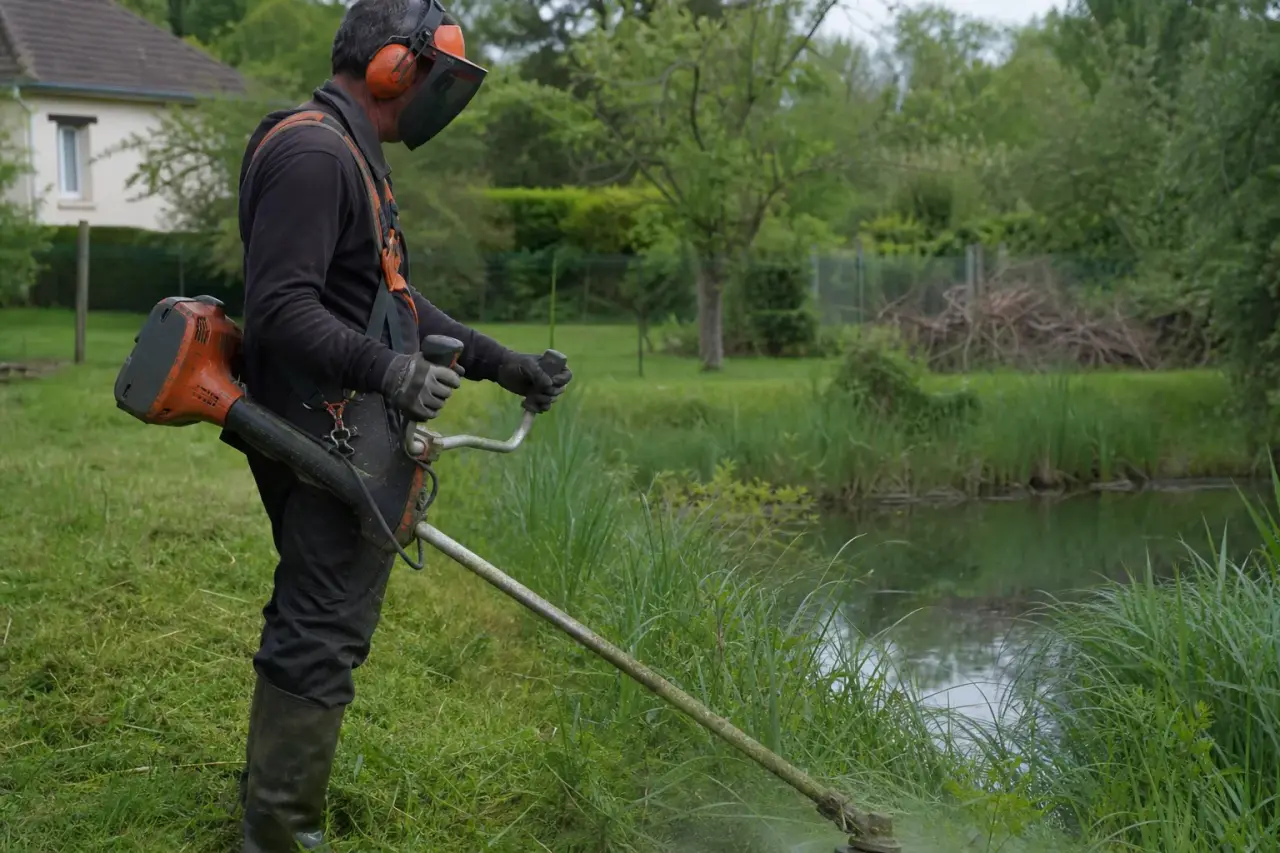 Gros plan d'un opérateur à la débroussailleuse coupant une strate herbacée dense au bord d'un fossé humide, équipé de protections, ambiance de plaine berrichonne.